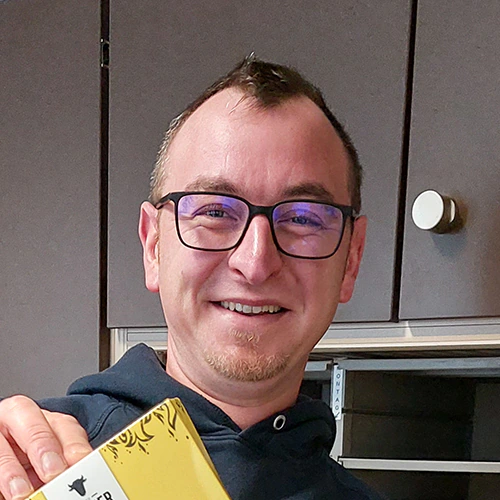 Porträt eines lächelnden Mannes mit Brille und dunklem Hoodie, der eine gelbe Verpackung in der Hand hält, vor einem Küchenschrank-Hintergrund.
